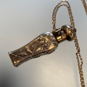 May include: A silver-colored perfume bottle pendant with an ornate floral design, suspended from a delicate gold-colored chain. The bottle has a rounded top and a long, slender body. The floral pattern is detailed and covers the entire surface.
