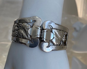 Puede incluir: Un brazalete de plata con un diseño de vid. El brazalete tiene bisagras y un patrón floral detallado.