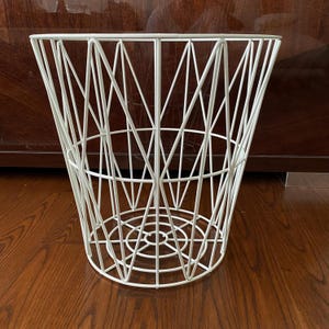 May include: A white wire basket with a geometric design. The basket has a conical shape, with a circular base and a series of intersecting lines forming the sides. The basket is made of metal and has a clean, modern aesthetic.