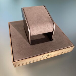 May include: A brown suede watch display stand with a black base. The stand has a rectangular base with the word 'Cartier' engraved on it. The stand is designed to hold a watch on the top.