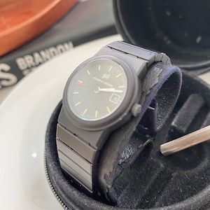 May include: A black Porsche Design wristwatch with a black leather band and a black face. The watch is in a black leather case.