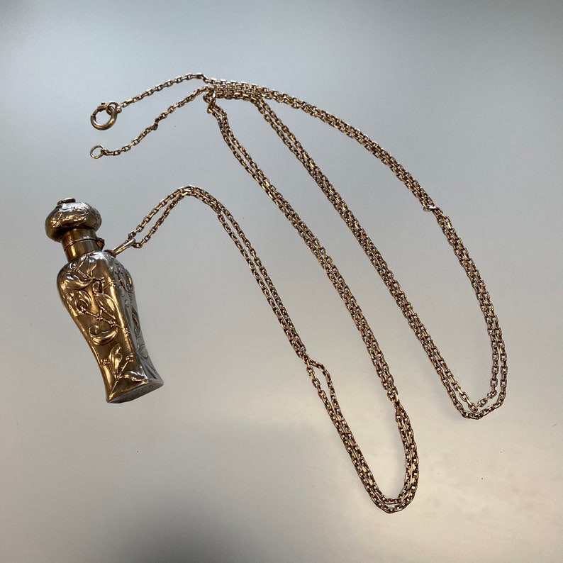 May include: A silver-toned perfume bottle pendant with an ornate floral design. The bottle is suspended from a delicate gold-colored chain. The chain is approximately 30 inches long.