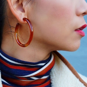May include: A woman wearing a white fleece jacket with brown trim and a blue and orange striped turtleneck sweater. She is wearing large brown hoop earrings.