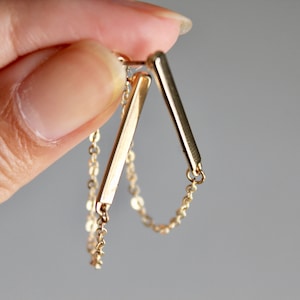 May include: Gold bar earrings with a delicate chain detail. The earrings are made of two thin gold bars connected by a chain.