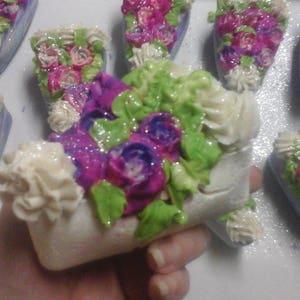 May include: Assortment of handmade soap bars shaped like cakes, decorated with colourful flowers and frosting. The soaps are in various shades of pink, purple, green, and white.