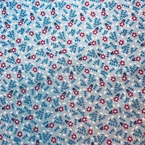 May include: Close-up of a fabric with a repeating floral pattern. The design features small, red flowers and dark blue leaves on a light blue background. Tiny white dots fill the spaces between the floral elements.
