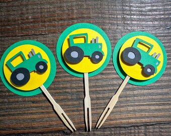 May include: Three green and yellow cupcake toppers featuring a green tractor design. The tractors have black wheels and are on a yellow circle with a green outer circle.