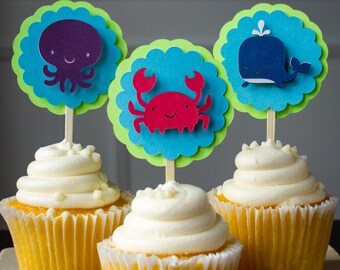 Puede incluir: Tres cupcakes decorados con adornos de papel de colores que representan un pulpo, un cangrejo y una ballena. Los cupcakes están cubiertos con glaseado blanco y adornados con chispas blancas.