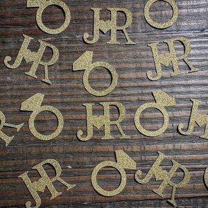 May include: Gold glitter ring and initial cutouts scattered on a wooden surface. The letters "J" and "R" are paired together. The ring cutouts have a diamond shape. Ideal for wedding decorations.