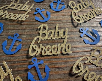 Puede incluir: Recortes de papel dorado con purpurina de las palabras "Baby Greene" con recortes de papel azul de anclas sobre un fondo de madera.