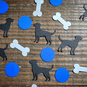 May include: A wooden surface scattered with black dog silhouettes, light blue bone shapes, and blue circular cutouts. The dog shapes are in various poses, and the elements are arranged in a playful pattern.