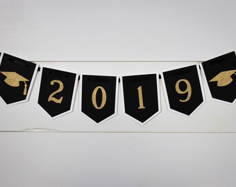 Pode incluir: Faixa de formatura preta e dourada com o ano de 2019 e gorros de formatura.