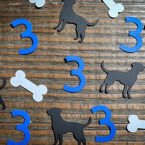 May include: A collection of paper cutouts including black dog silhouettes, blue number 3s, and white bone shapes. The cutouts are scattered on a wooden surface, creating a playful and decorative arrangement.