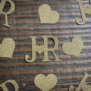 May include: Gold glitter letters and heart shapes scattered on a wooden surface. The letters include "J", "R", and "M". The hearts and letters are likely used for crafting or decoration, possibly for a wedding or special event.