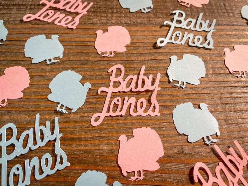 K&ouml;nnte beinhalten: Rosa und blaue Papierschnitte von Truth&auml;hnen und dem Text "Baby Jones" auf einem Holzgrund.