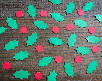 May include: Green paper holly leaves and red paper circles scattered on a brown wooden surface.