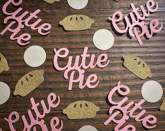 May include: Pink "Cutie Pie" script lettering and gold glitter pie slice cutouts are scattered on a wooden surface, along with white circular confetti. The image has a warm, inviting aesthetic, perfect for a baby shower or birthday.