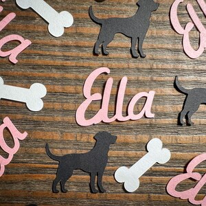 May include: A collection of cut-out shapes on a wooden surface. The shapes include the name "Ella" in pink script, black dog silhouettes, and white bone shapes. The arrangement is scattered across the wood grain background.