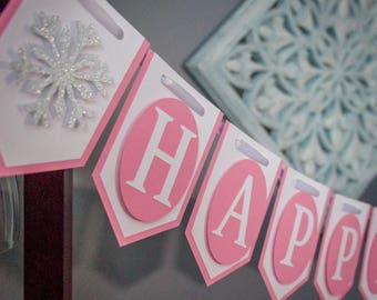 May include: A pink and white banner with the word "HAPPY" in white letters on pink circles. Each letter is on a separate pennant. The banner is decorated with a white snowflake.