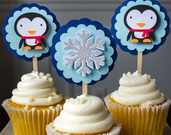 May include: Three cupcakes decorated with blue and white snowflake, penguin, and penguin cupcake toppers. The cupcakes are frosted with white frosting and topped with white sprinkles.