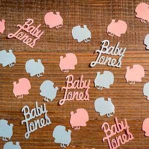 K&ouml;nnte beinhalten: Rosa und blaue Papier-Truthahn-Ausschnitte mit dem Text "Baby Jones" auf einer Holzoberfl&auml;che verstreut.