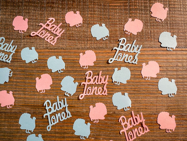 K&ouml;nnte beinhalten: Rosa und blaue Papier-Truthahn-Ausschnitte mit dem Text "Baby Jones" auf einer Holzoberfl&auml;che verstreut.