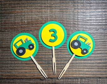 May include: Three green and yellow cupcake toppers featuring a tractor design and the number 3. The toppers are on wooden sticks.