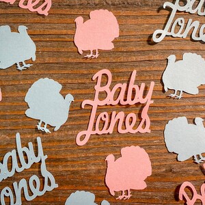 K&ouml;nnte beinhalten: Rosa und blaue Papierschnitte von Truth&auml;hnen und dem Text "Baby Jones" auf einem Holzgrund.
