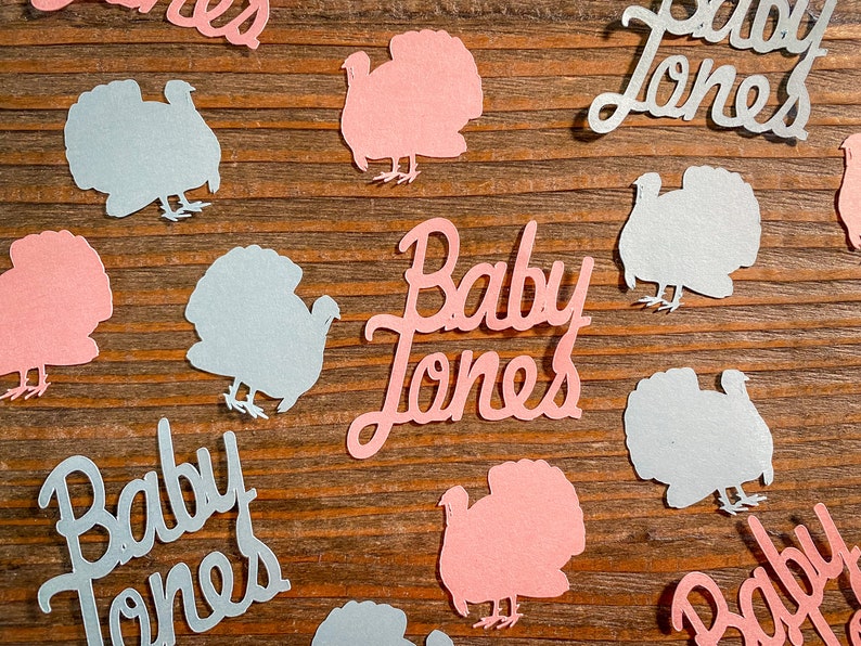 K&ouml;nnte beinhalten: Rosa und blaue Papierschnitte von Truth&auml;hnen und dem Text "Baby Jones" auf einem Holzgrund.
