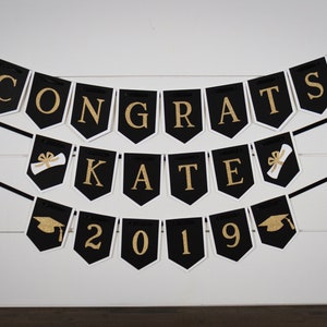 May include: Black and gold graduation banner with the text "Congrats Kate 2019". The banner features graduation caps and diplomas.