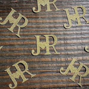 Può includere: Ritagli di lettere JR con glitter dorati sparsi su una superficie di legno. Le lettere sono in un carattere script e sono ideali per decorazioni di matrimoni o feste. I glitter aggiungono un tocco festoso.