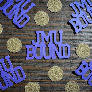 May include: Purple cut-out letters spelling "JMU BOUND" and scattered gold glitter circles on a dark wood grain surface. The letters are in a bold, sans-serif font. The overall theme suggests a celebratory or decorative item.
