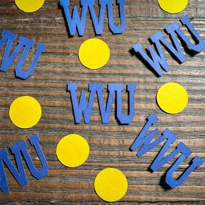 May include: Blue cut-out letters spelling "WVU" are scattered across a wooden surface, interspersed with yellow circular cutouts. The letters and circles are arranged in a visually appealing pattern, creating a decorative effect.