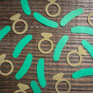 Op de afbeelding: Gouden glitter ring uitsparingen en groene gebogen vormen zijn verspreid op een houten oppervlak. De ring uitsparingen hebben een diamantvormig accent. De groene vormen zijn gebogen en langwerpig.