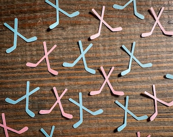 May include: A wooden surface with scattered paper cutouts of hockey sticks in blue and pink. The sticks are crossed at the top and the bottom.