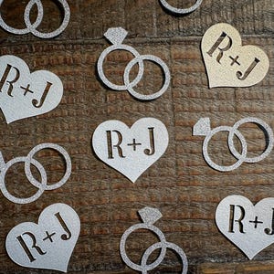 May include: Scattered silver glitter wedding ring and heart-shaped confetti with the initials "R+J" cut out. Some rings have a diamond accent. The confetti is on a dark brown wooden surface.