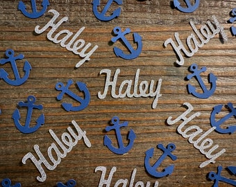Puede incluir: Confetti de purpurina plateado con el nombre "Haley" y confetti azul de ancla. El confetti está esparcido sobre una superficie de madera.