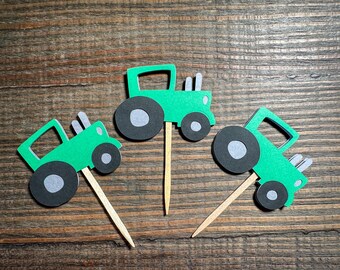 May include: Three green and black tractor cupcake toppers with wooden sticks. The tractors have black wheels and are made of paper.