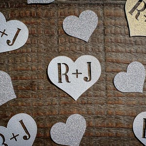 May include: Scattered heart-shaped confetti with glittery silver and gold finishes. Each heart features the initials "R+J" cut out. The confetti is arranged on a textured, brown wooden surface, ideal for weddings or celebrations.