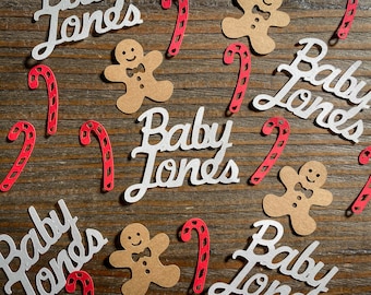 Può includere: Sagome di carta bianca di bastoncini di zucchero e omini di pan di zenzero con il testo "Baby Jones" ripetuto su diverse sagome. Le sagome sono sparse su una superficie di legno marrone.