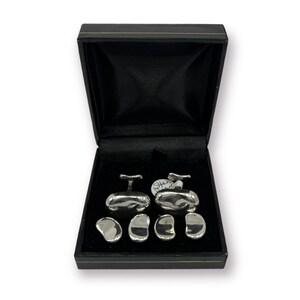 May include: A set of silver cufflinks and shirt studs presented in a black box. The cufflinks have a unique, organic shape, and the shirt studs are small, matching the cufflinks. The box is open, revealing the contents against a black velvet lining.