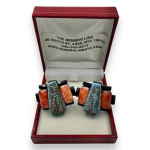 May include: A pair of cufflinks in a red box. The cufflinks are made of black and silver metal and feature a colorful design of an Egyptian woman with hieroglyphics. The box has a label that reads "The Missing Link, 40 W 25th St, #222, NYC 10010, 646-345-8514, www.missinglinknyc.com".