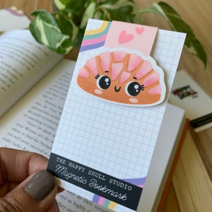 May include: A pink and brown cartoon croissant with a happy face and big eyes. The bookmark is magnetic and has a grid pattern on the back. The text "THE HAPPY SKULL STUDIO Magnetic Bookmark" is printed on the bottom of the bookmark.