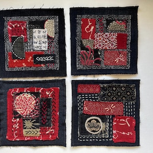 May include: Four square fabric coasters featuring a boro-inspired patchwork design. Each coaster is constructed from dark navy blue fabric with various red, black, and grey textile patches, accented by white hand-stitched embroidery and Japanese kanji.
