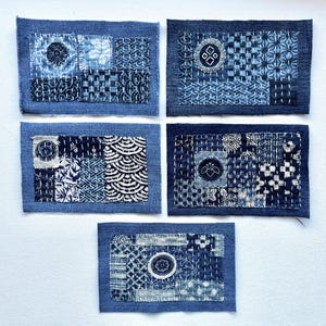 May include: Five indigo blue textile art pieces with various geometric and floral patterns. Each piece is rectangular with a denim border and features hand-stitched details in white thread. The designs include circles, squares, and wave-like motifs.