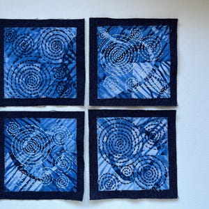 May include: Four square fabric coasters with a blue and white spiral design. Each coaster has a dark blue border and a unique pattern of concentric circles stitched with white thread. The coasters are arranged in a square.