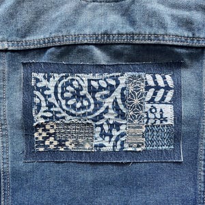 May include: Close-up of a denim jacket back with a rectangular patch. The patch is made of various blue and white patterned fabrics, stitched together with white thread. The jacket is a medium wash denim.
