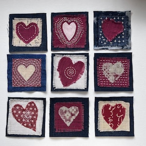 May include: Nine square fabric patches arranged in a grid, each featuring a heart design. The patches use dark blue denim backgrounds with various heart shapes in shades of red, pink, and cream, accented with visible hand-stitched embroidery.