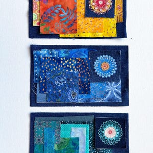 May include: Three rectangular fabric collages featuring layered patches of various textiles. Each piece includes a dark blue denim base, assorted patterned fabric squares in warm, cool, and teal tones, and a central embroidered floral motif.