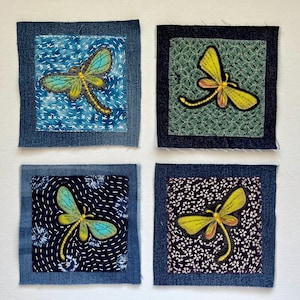 May include: Four square fabric patches with dragonfly designs. Each patch features a denim border and a central design of a dragonfly in shades of yellow, blue, and black. The backgrounds vary with patterns of blue, green, and white.